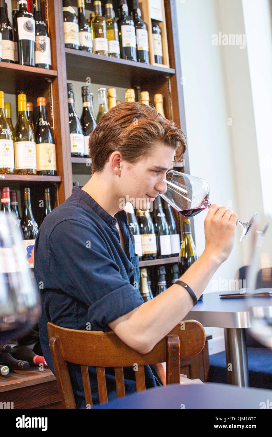 Giovane maschio odore sapore di vino rosso in ristorante sullo sfondo di mensole con bottiglie. Uomo degustazione un bicchiere di vino rosso. Foto Stock