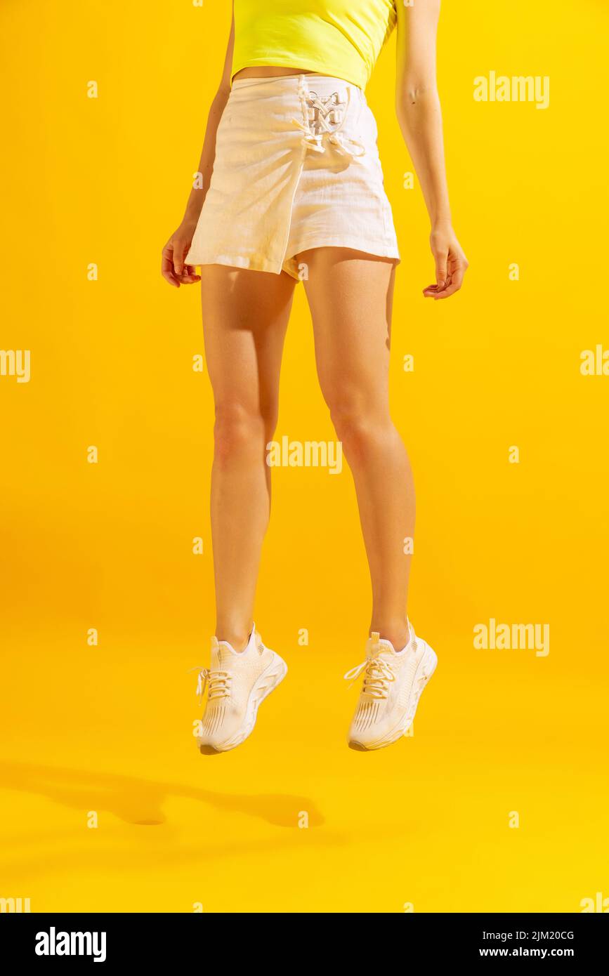 Gambe femminili sottili con gonna corta bianca e sneakers che si sollevano sopra il pavimento isolato su sfondo giallo. Concetto di bellezza, arte, moda, levitazione Foto Stock