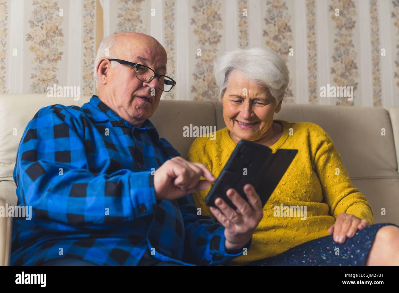 Uomo anziano caucasico seduto con la moglie su un divano nel soggiorno e mostrando le sue foto sul suo smartphone. Foto di alta qualità Foto Stock