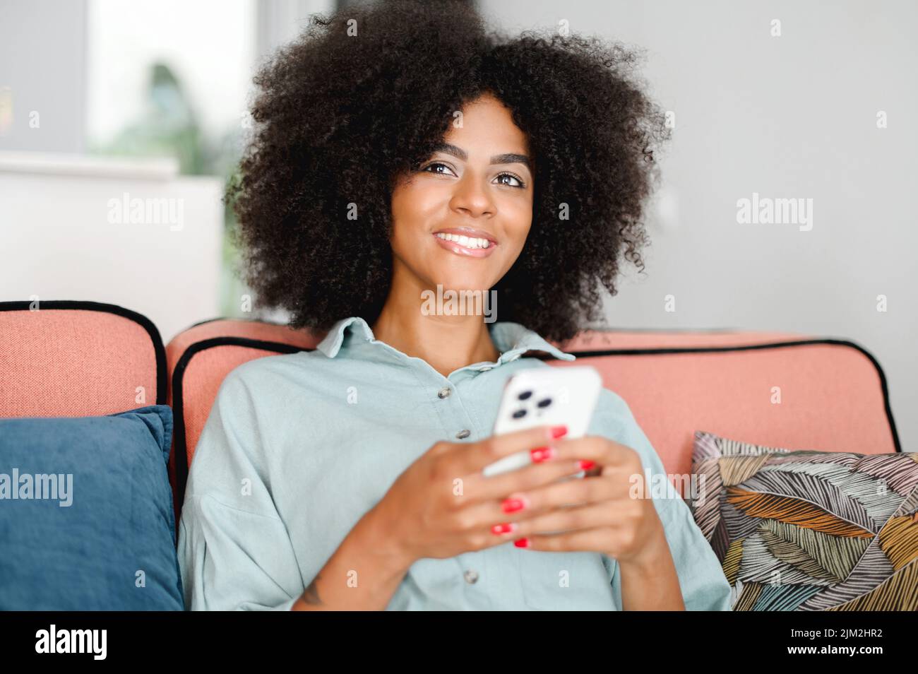 Giovane donna africana sorridente seduta sul divano a casa e con smartphone, SMS, utilizzando smartphone per lo shopping online. Tecnologie e concetto di persone Foto Stock