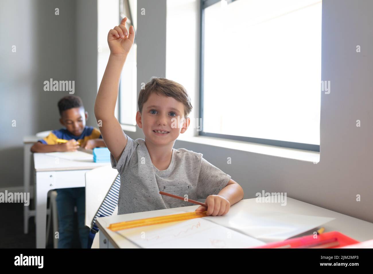 Ragazzo elementare caucasico sorridente con mano sollevata seduto alla scrivania in classe Foto Stock