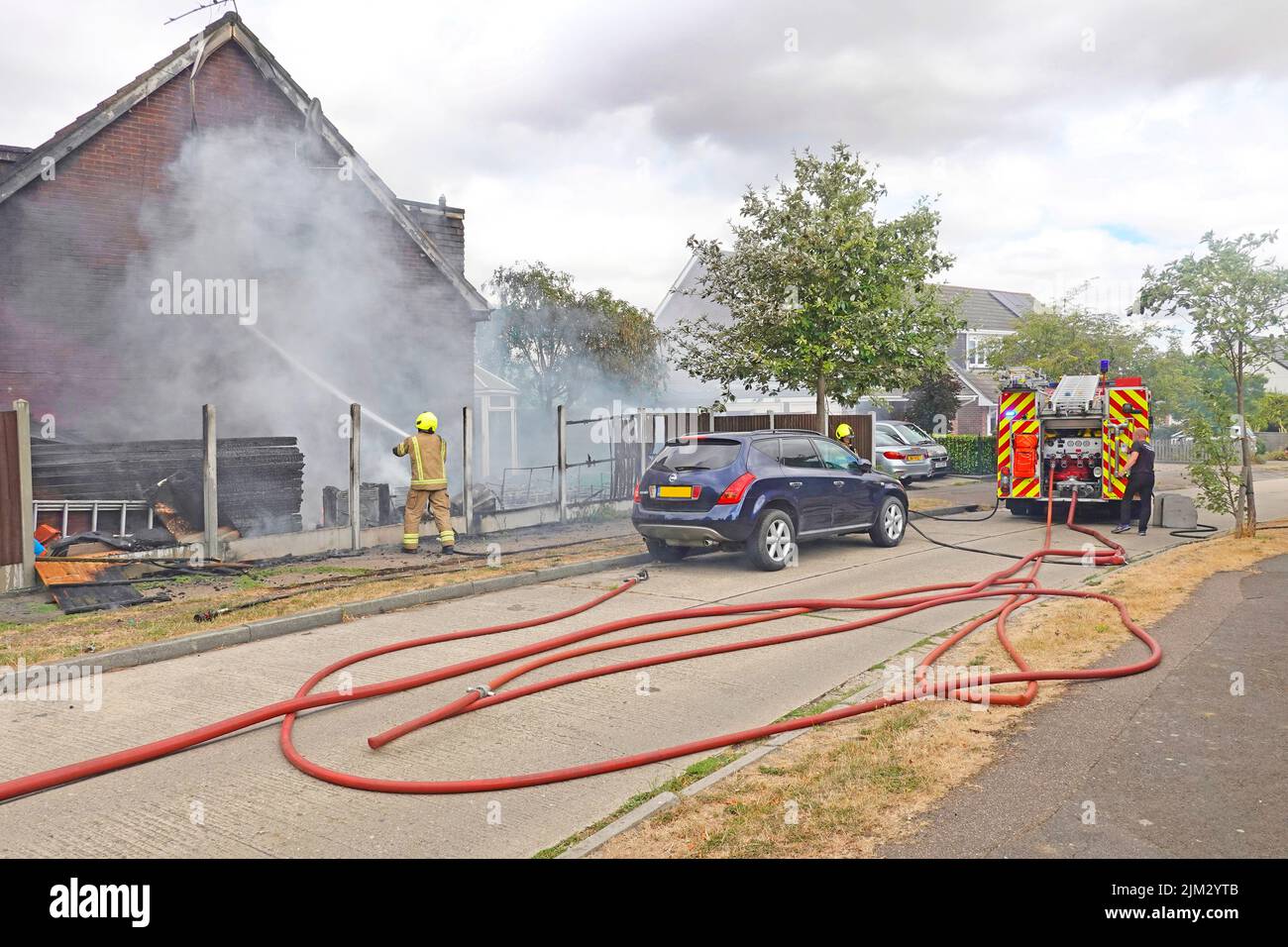 Pompiere che porta casa fuoco Blaze sotto controllo tubi acqua su Essex Fire & Rescue Service fuoco motore tender & resti di legno recinto Inghilterra UK Foto Stock