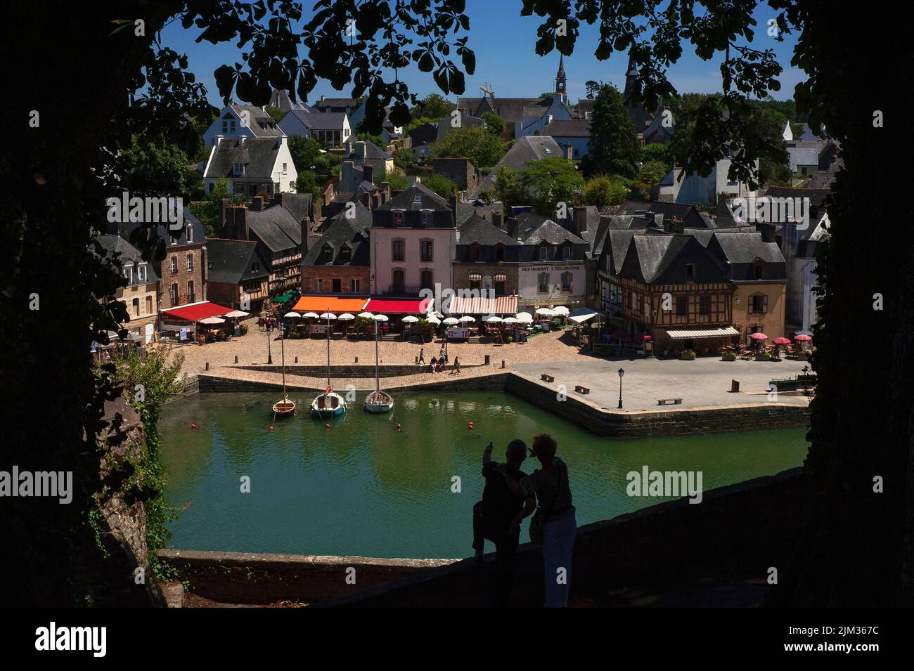 Una coppia si staglia contro il verde Loch stand Loch guardando e puntando verso il basso a Place Saint-Sauveur nel quartiere Saint-Goustan di Auray, Morbihan, Bretagna meridionale, Francia, fiancheggiata da ristoranti e caffè marciapiede con tende colorate e ombrelloni. Saint-Goustan, un tempo un porto di pesca e commerciale, è ora una popolare destinazione turistica che onora una visita del 1776 da parte del padre fondatore degli Stati Uniti, Benjamin Franklin. Foto Stock