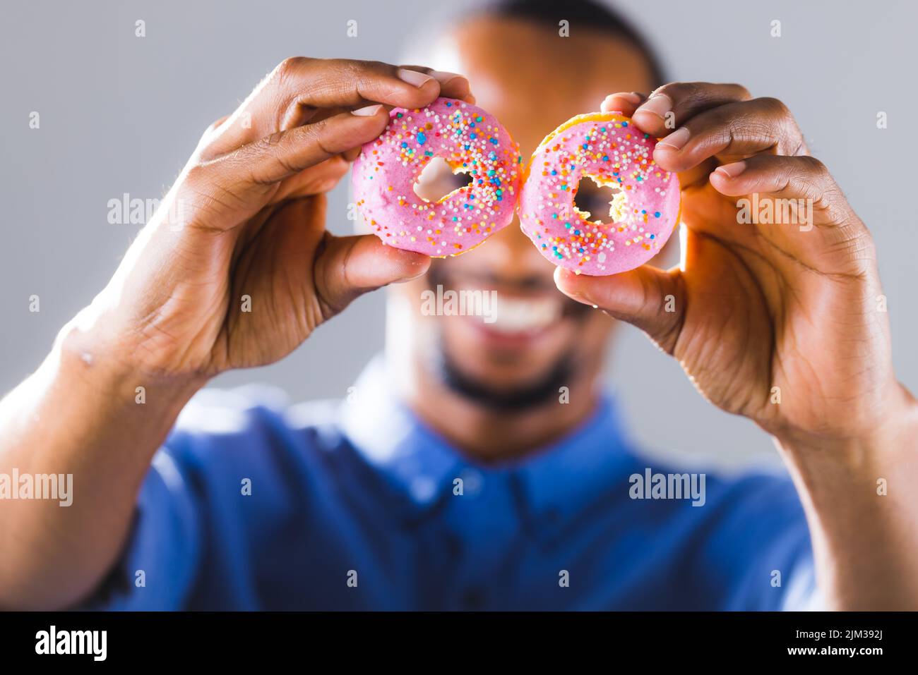 Uomo afroamericano sorridente che tiene le ciambelle rosa fresche davanti al viso contro lo sfondo grigio Foto Stock