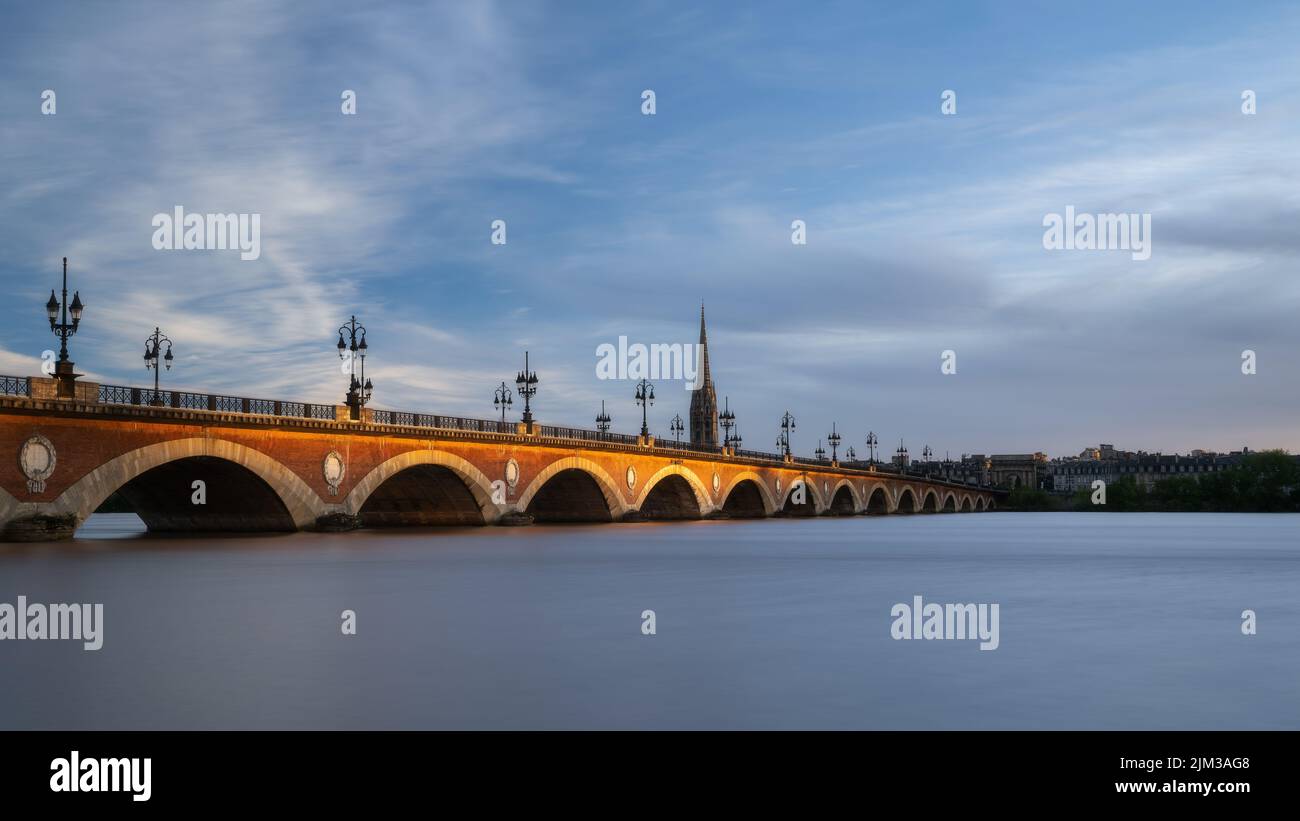 Pont de Pierre che attraversa il fiume Garonna nella città di Bordeaux illuminata dal sole tramonto con la Basilica Saint-Michel in background Foto Stock
