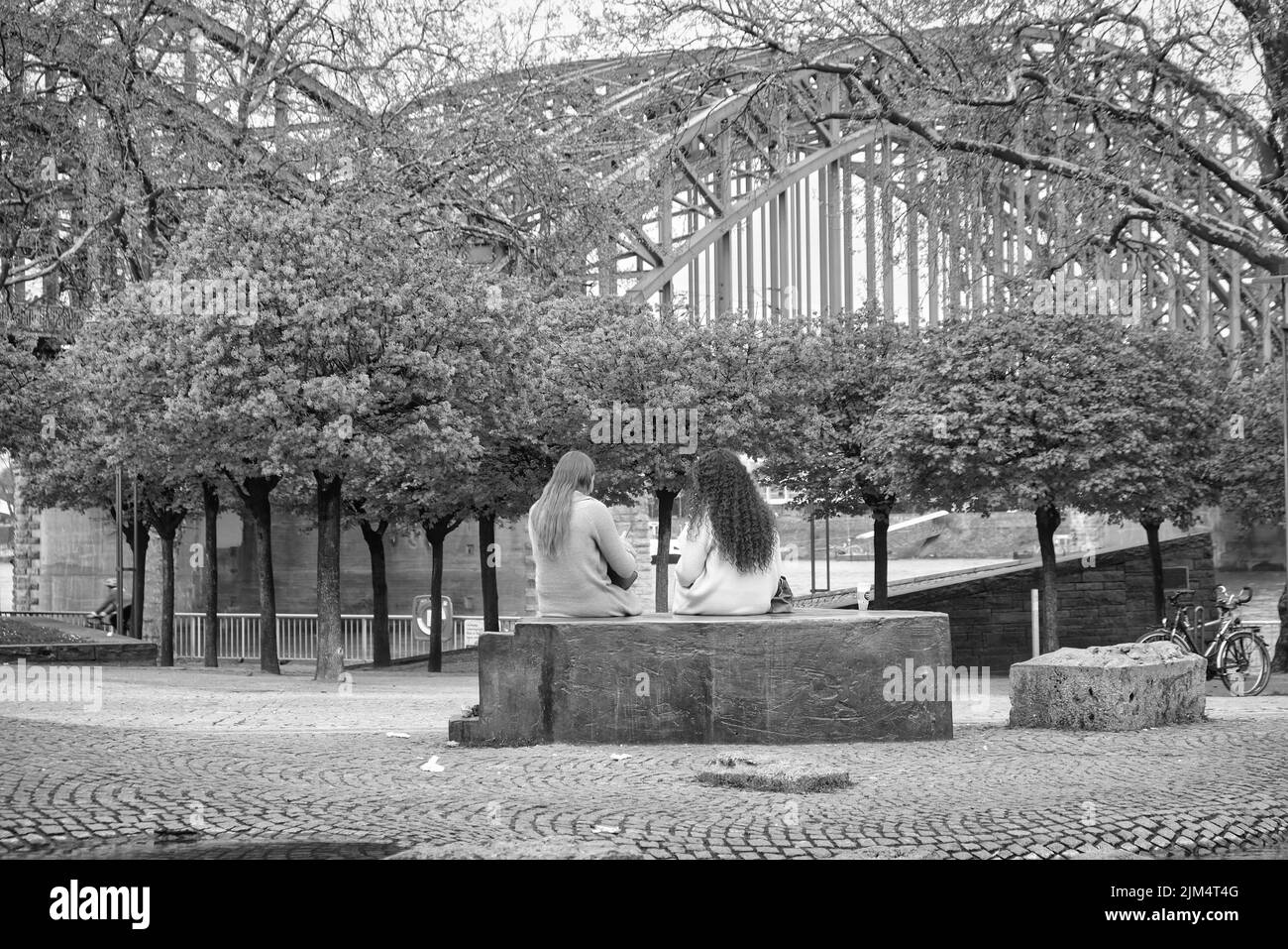 Due giovani donne sedute sulle rive del Reno a Colonia, Germania, ponte sullo sfondo, foto di strada in bianco e nero Foto Stock