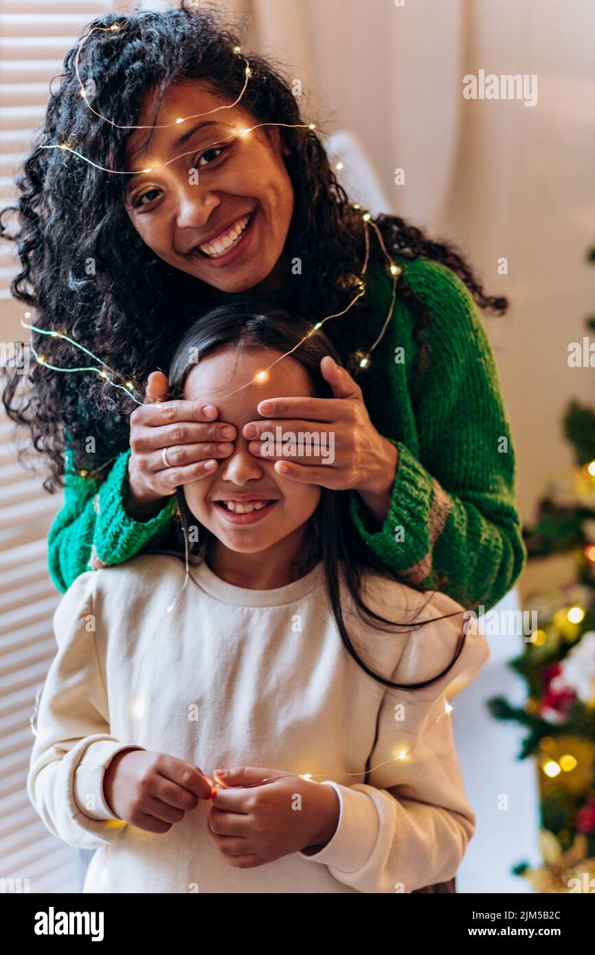 Bruna scolgirl e curly afroamericana madre posare gli occhi chiudendo con le mani in garland illuminante e sorridere in elegante soggiorno Foto Stock