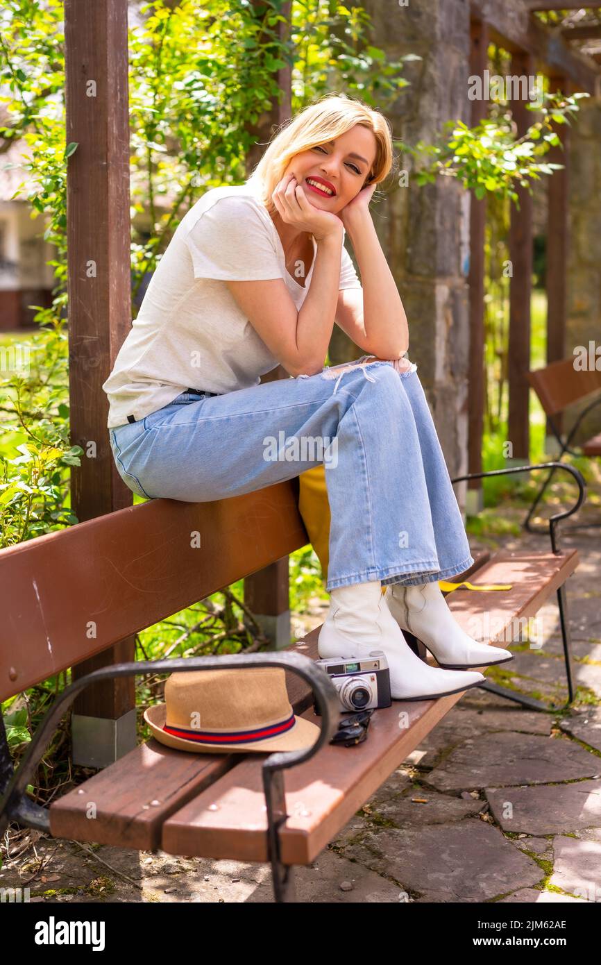 Una ragazza caucasica bionda in un top bianco e jeans seduti su una panca nel parco con una macchina fotografica, cappello e zaino accanto a lei Foto Stock