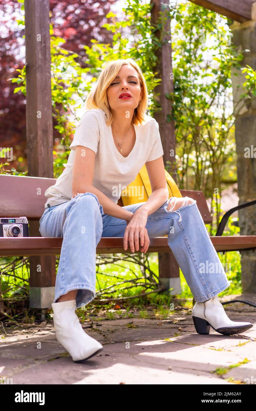 Un colpo verticale di una ragazza caucasica bionda in cima bianca e jeans seduti su una panca nel parco Foto Stock