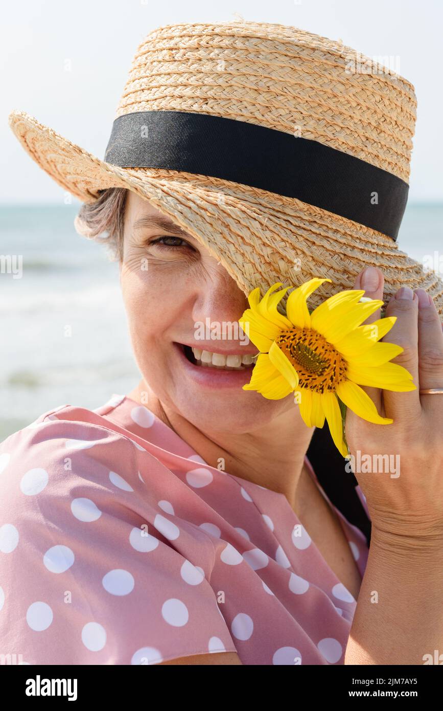 Donna sposata in abito rosa con pallini bianchi allegri e giocosamente sorridenti alla macchina fotografica che tiene cappello di paglia e girasole, su sfondo mare sfocato Foto Stock