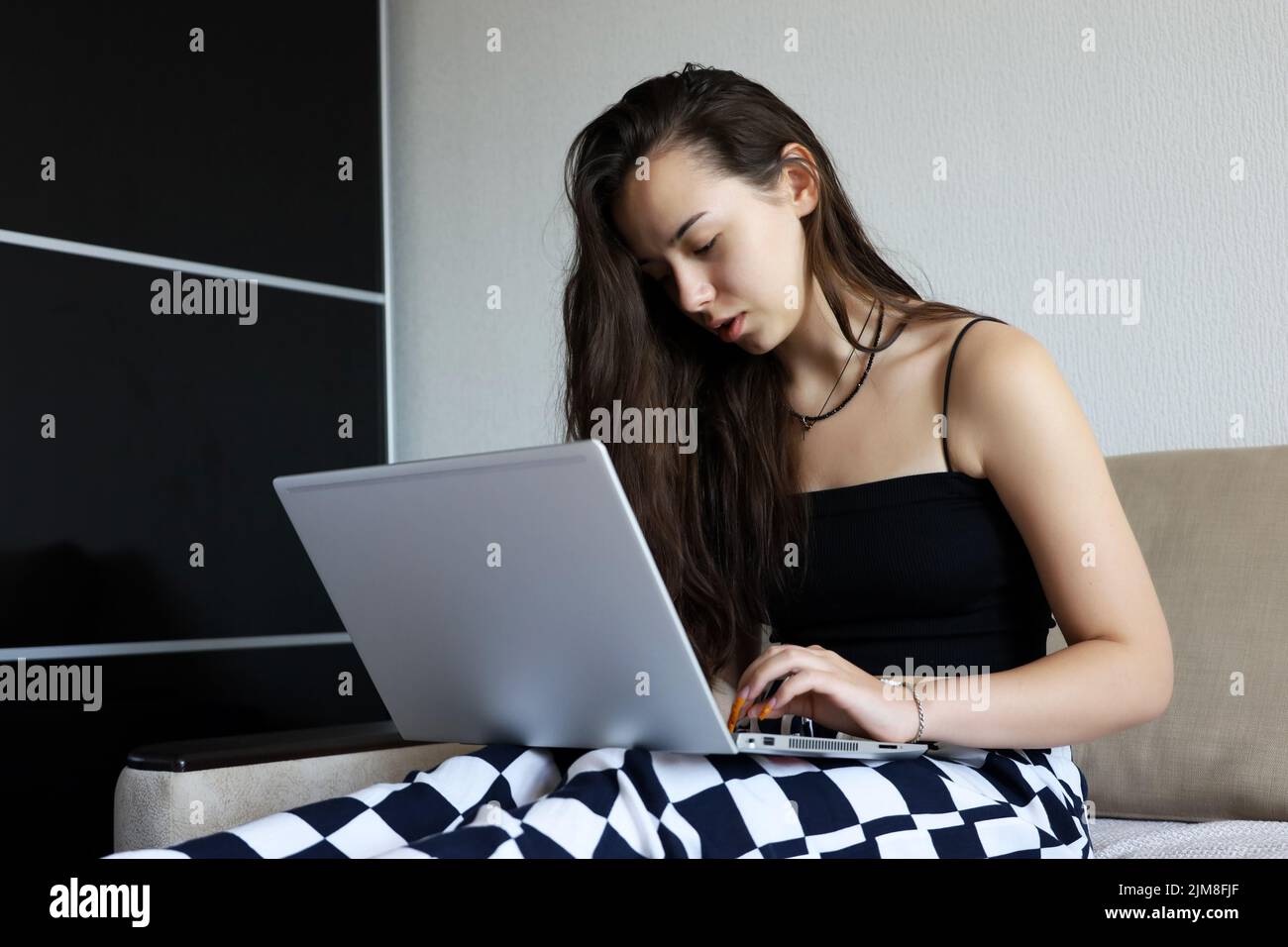 Bella ragazza giovane con capelli lunghi che indossa la parte superiore nera seduta con il computer portatile sul divano. Concetto di lavoro, studio o tempo libero a casa Foto Stock