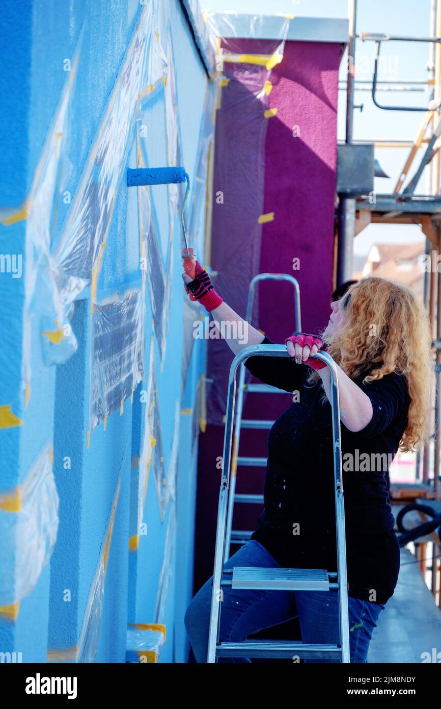 donna pittrice rossa matura per adulti con capelli ricci rossi e occhiali da sole, si alza sulla scala e dipinge colori poligonali artistici in vernice blu Foto Stock