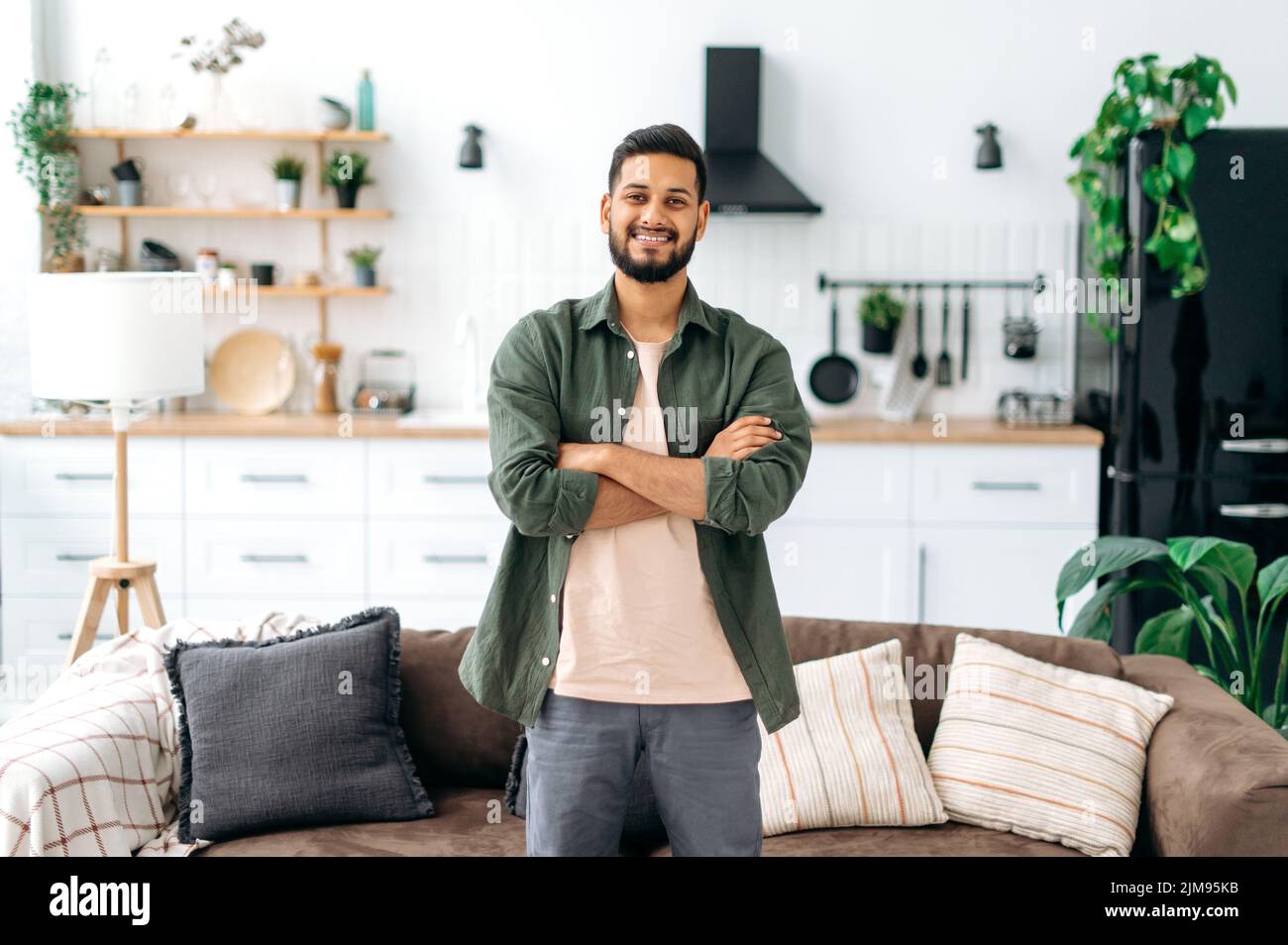 Ritratto di un bel ragazzo positivo indiano o arabo moderno, in abiti casual elegante, in piedi a casa nel soggiorno sullo sfondo della cucina, con le braccia incrociate, guarda la macchina fotografica, sorriso Foto Stock