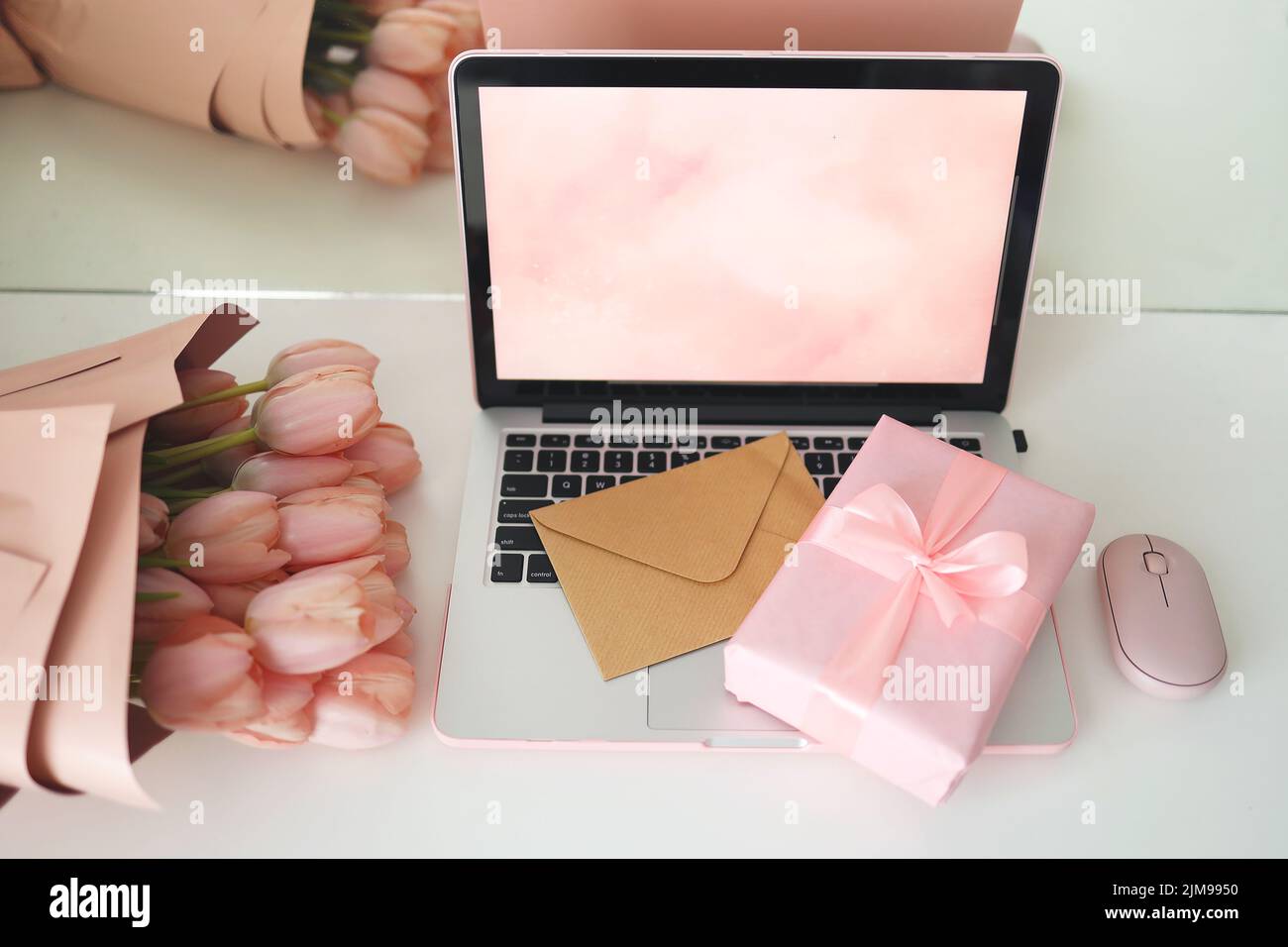 Elegante scrivania femminile con tulipani rosa, computer portatile e busta. Visualizzazione superiore dello schermo del notebook. Scatola regalo rosa e mouse rosa per computer. Foto Stock
