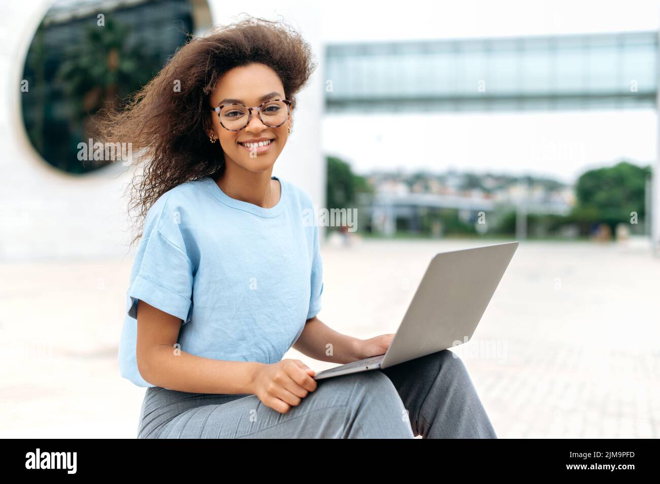 Ritratto di una giovane donna d'affari afroamericana elegante e moderna con occhiali, ceo azienda, freelance, studentessa femminile, seduta con laptop all'aperto, guardando la macchina fotografica, sorridente amichevole Foto Stock