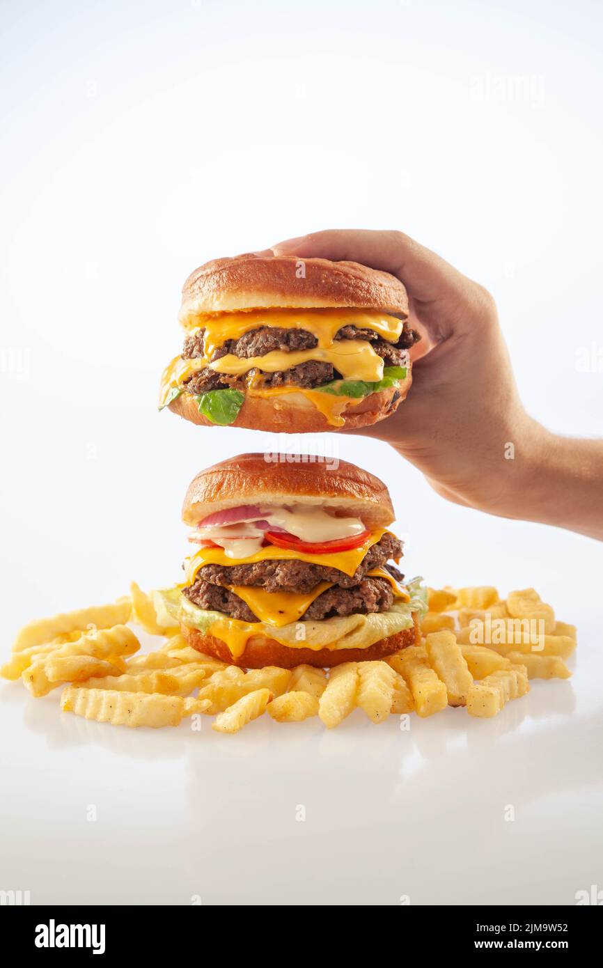 Mano che tiene un hamburger di formaggio gourmet sopra un altro hamburger di manzo con patatine sul fondo Foto Stock