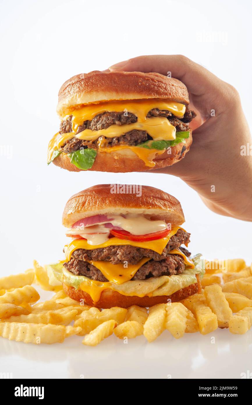 Mano che tiene un hamburger di formaggio gourmet sopra un altro hamburger di manzo con patatine sul fondo Foto Stock