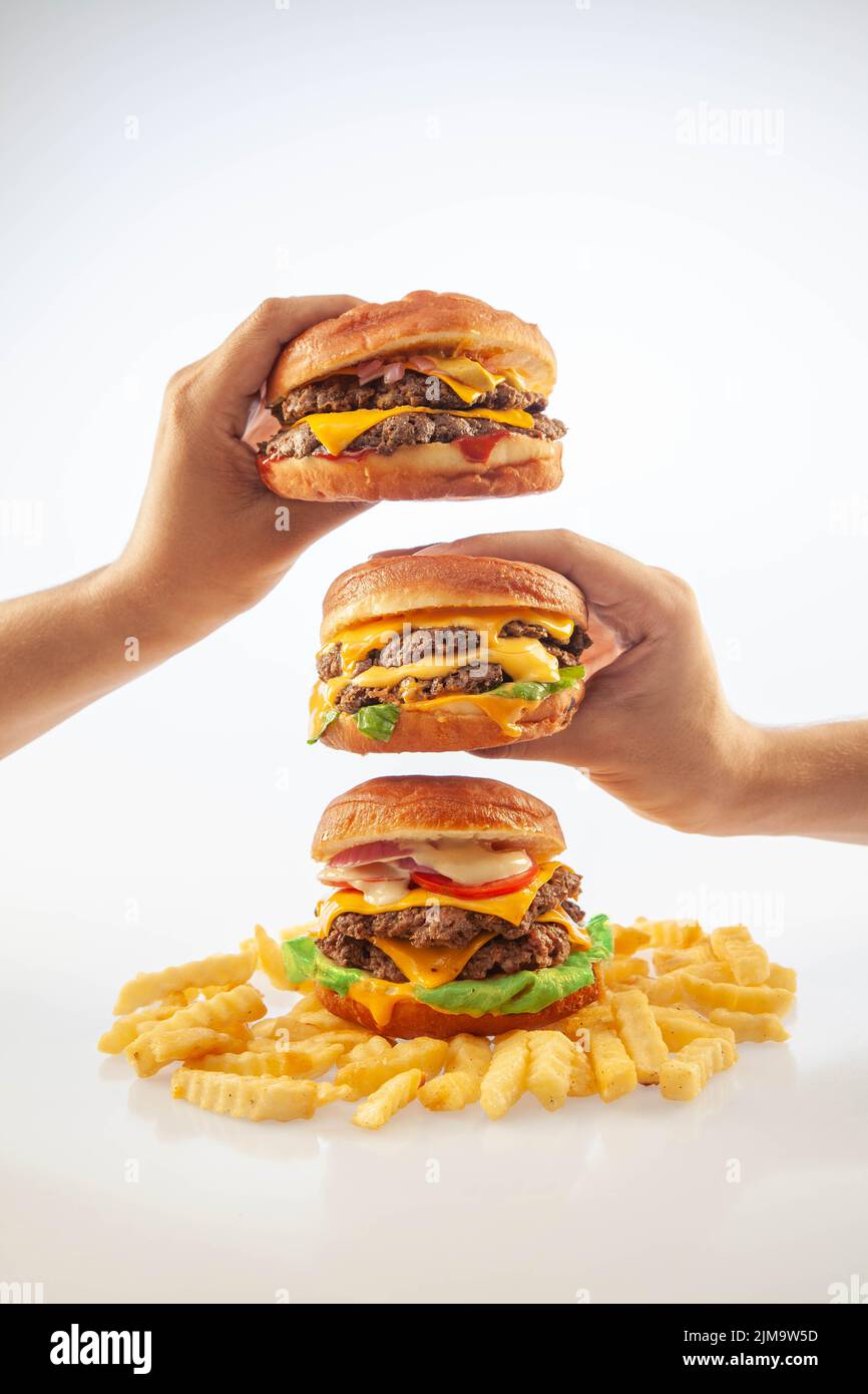 Due mani che tengono un hamburger di formaggio gourmet uno sopra l'altro con hamburger di manzo con patatine sul fondo Foto Stock