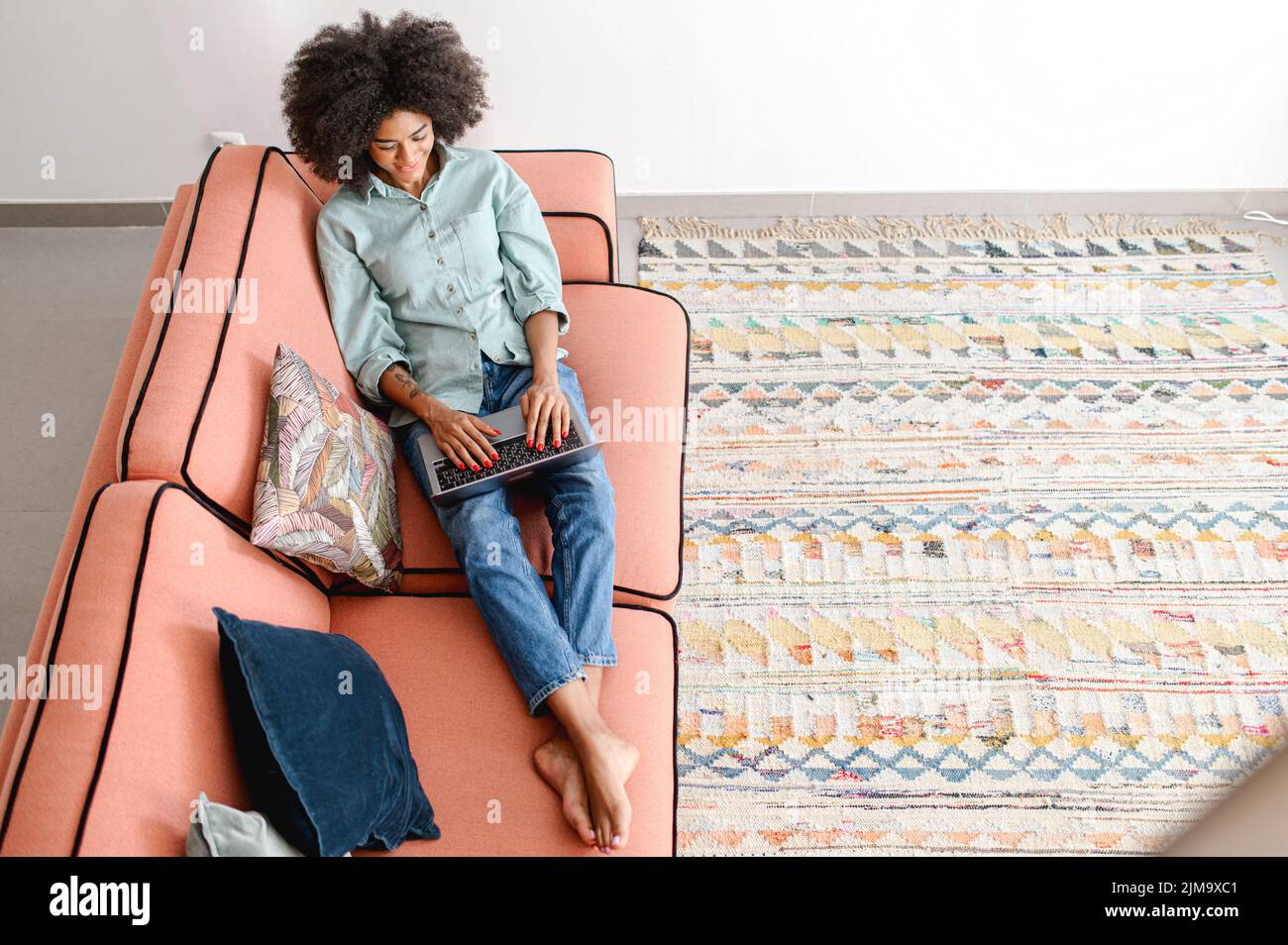 Vista ad alto angolo della donna afro-americana usando il computer portatile sdraiato su un comodo divano in appartamento moderno. Etnia femminile freelance digitando sulla tastiera, studiando o lavorando a distanza, vista dall'alto Foto Stock