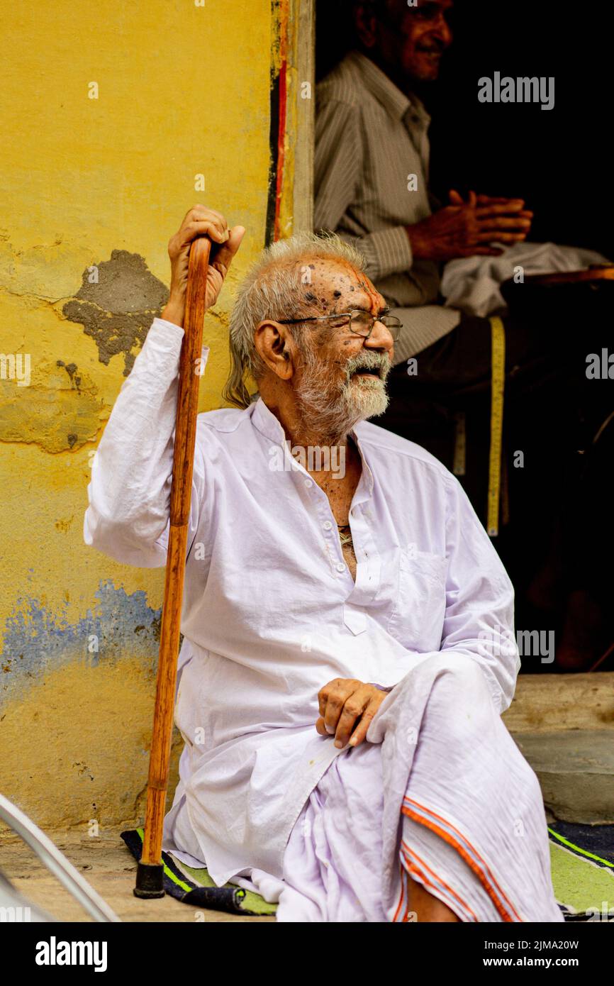 Un vecchio indiano in abiti bianchi tradizionali seduti contro un muro giallo, Nathdwara, India Foto Stock