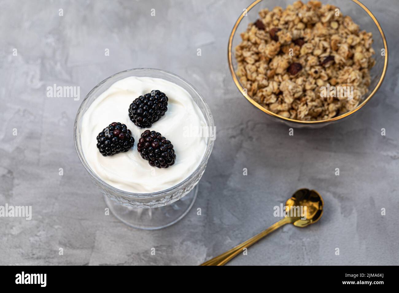 Dessert con yogurt, muesli fatti in casa e more fresche, serviti in vetro chiaro trendy con motivi geometrici e cucchiaio dorato, sfondo grigio. Sana colazione vegana o snack. Spazio di copia, messa a fuoco selettiva Foto Stock