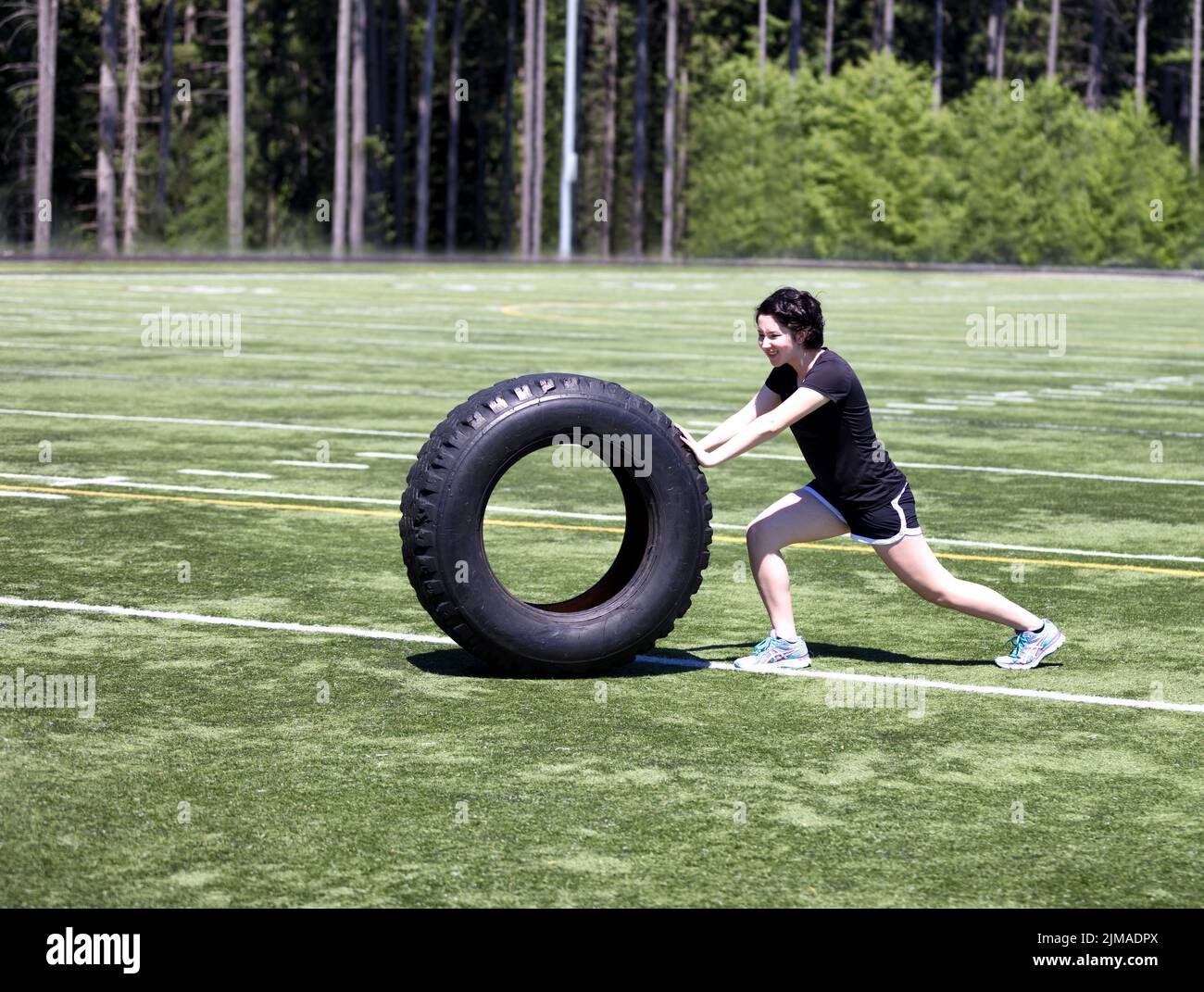 Ragazza teen che spinge la gomma pesante sul campo sportivo durante il giorno caldo Foto Stock