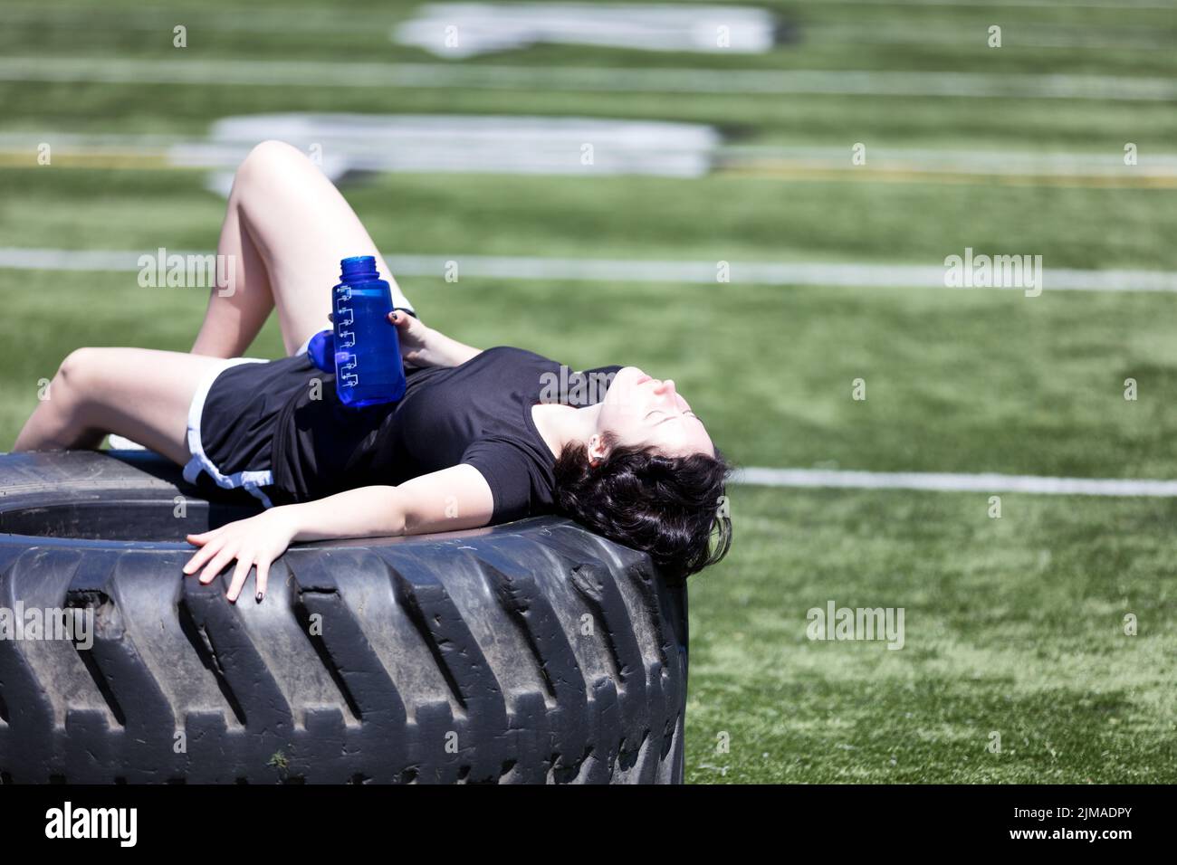 Ragazza adolescente esausta che riposa su pneumatico pesante del camion nel mezzo del campo sportivo durante il giorno caldo Foto Stock