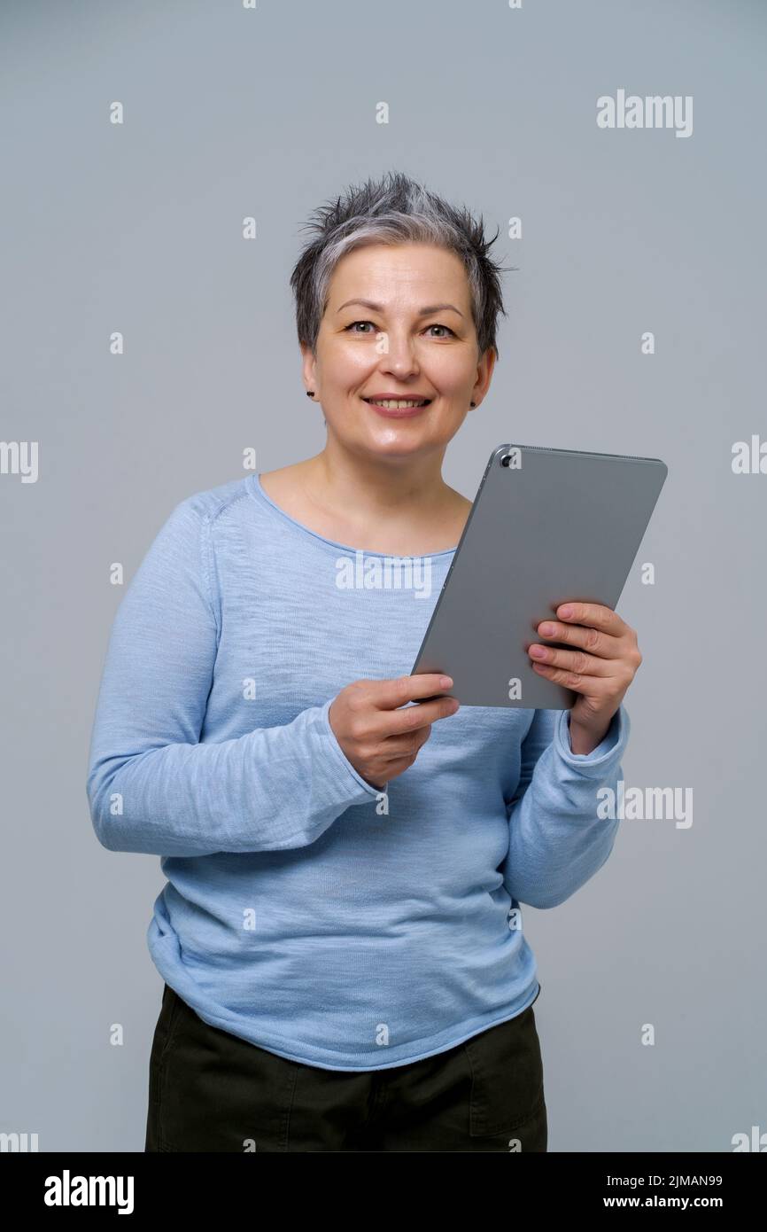Donna grigia matura con capelli che tiene un tablet digitale in funzione o che fa shopping online, controllando i social media. Bella donna in blusa blu isolato su sfondo bianco. Foto Stock