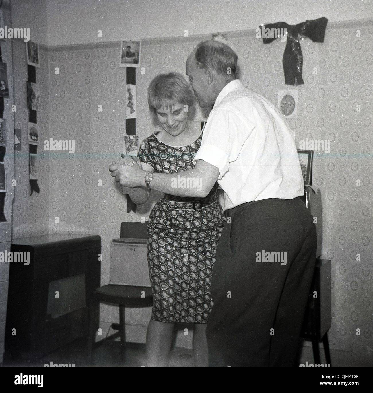 1960s, storico, un Signore di mezza età e una giovane donna che hanno una danza insieme al chiuso in una festa di casa, Inghilterra, Regno Unito, biglietti d'auguri montati sul muro. Un armadio radio dell'epoca si trova all'angolo. Foto Stock