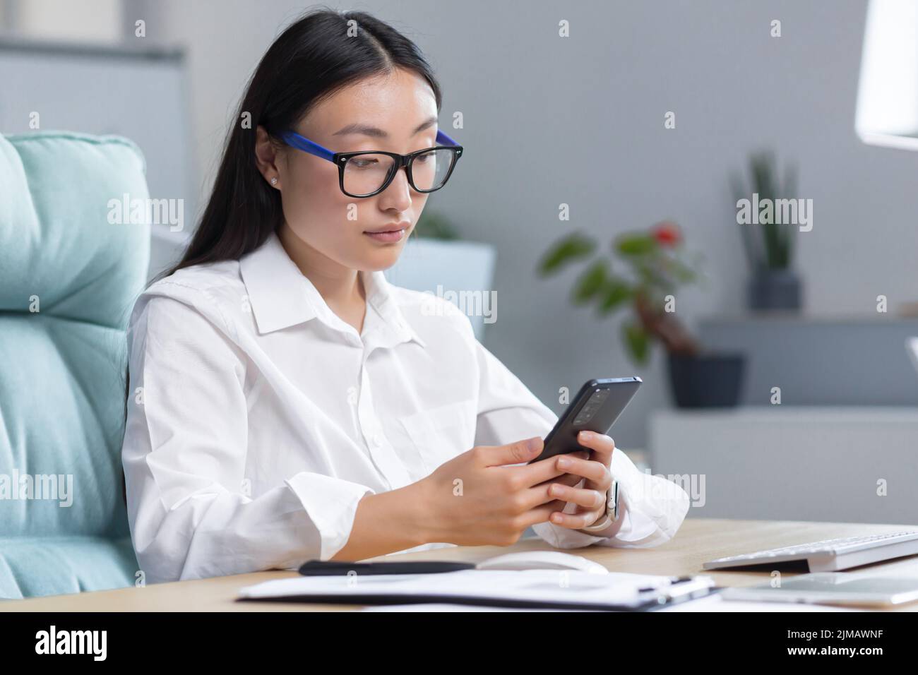 Una giovane bella donna asiatica d'affari in occhiali sta tenendo un telefono nelle sue mani, componendo un messaggio. Seduta su una scrivania in un ufficio moderno. Foto Stock