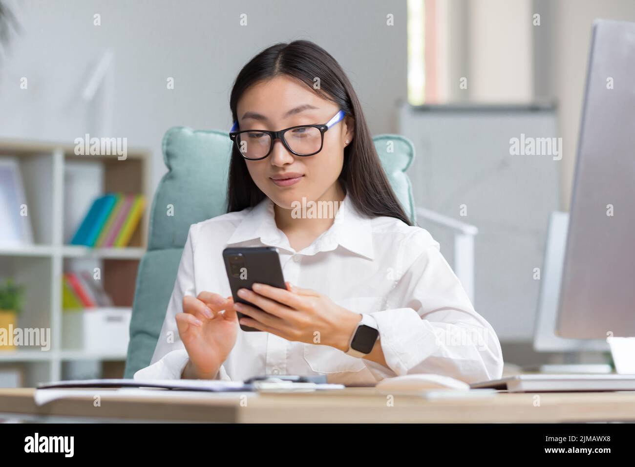 Una giovane bella donna asiatica d'affari in occhiali sta tenendo un telefono nelle sue mani, componendo un messaggio. Seduto su una scrivania in un ufficio moderno, guardando la fotocamera Foto Stock