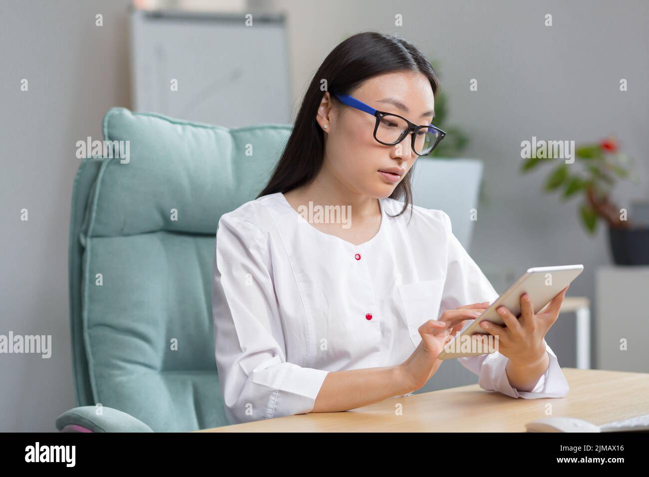 Ammissione online all'ospedale. Una giovane bella donna asiatica medico registra online la storia medica di un paziente in un tablet. Digitazione in un tablet. Seduta al tavolo nell'ufficio di avvolgimento. Foto Stock