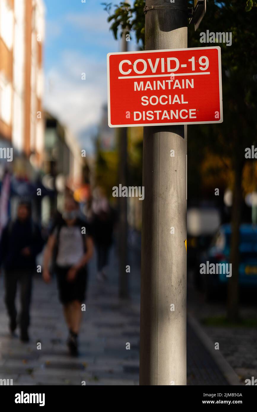Uno scatto verticale di un cartello rosso e bianco "COVID-19 Maintain Social Distance" sul lampione con i pedoni sullo sfondo sfocato Foto Stock