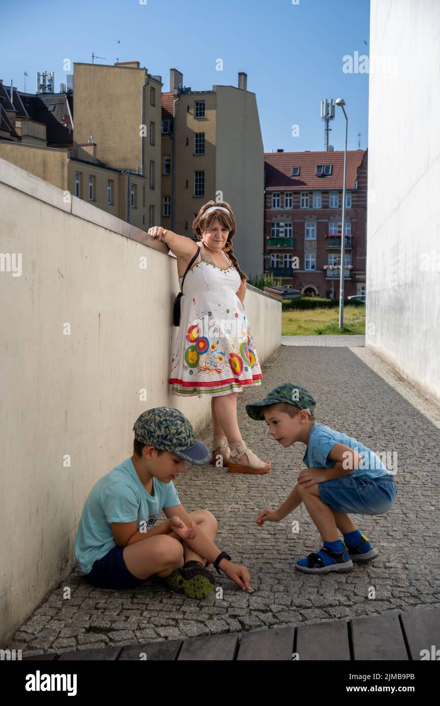 Una donna anziana che guarda due ragazzi in ginocchio e che siede a terra di fronte all'ingresso del museo Brama Poznania. Foto Stock