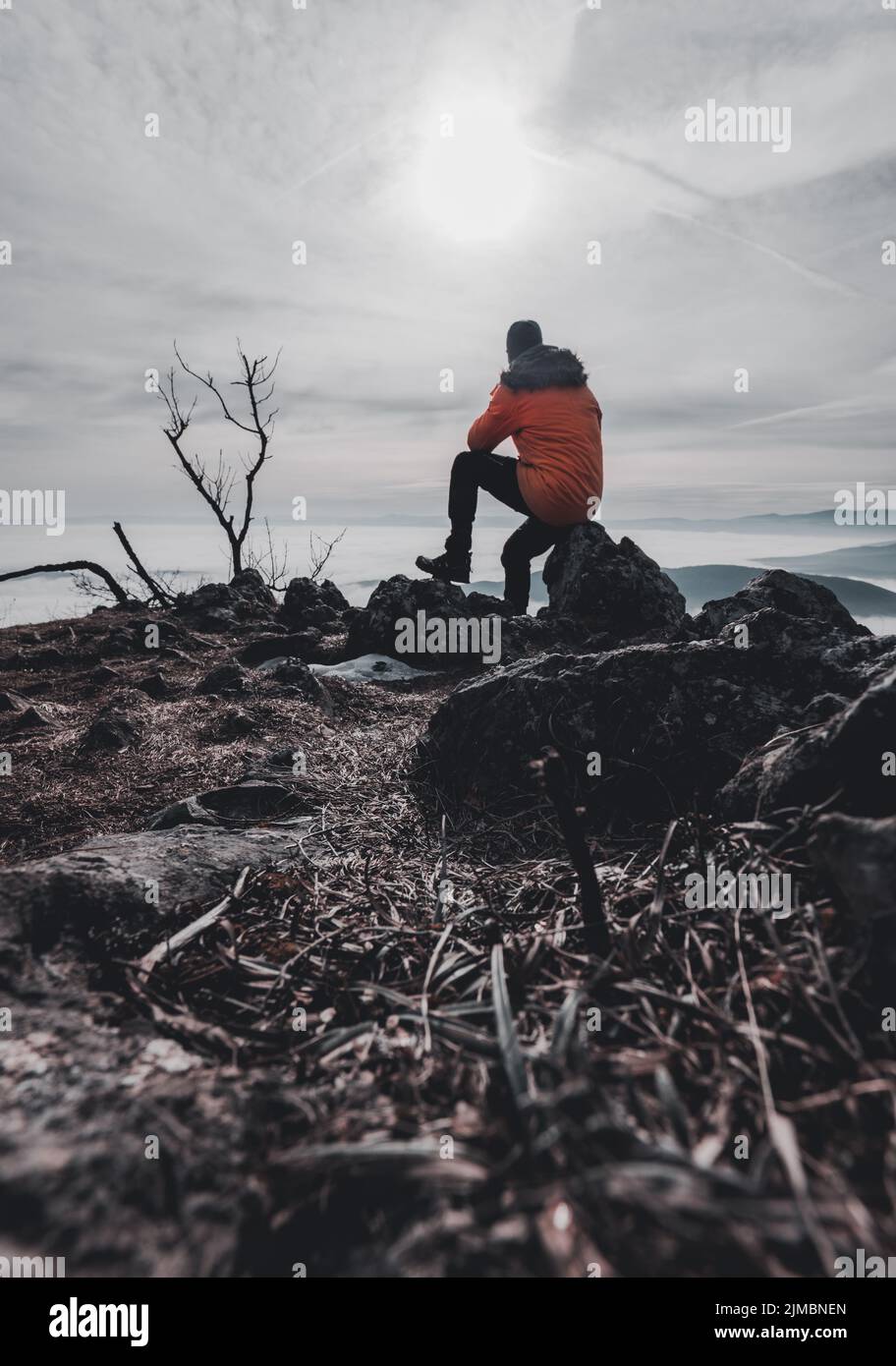 Vista posteriore di un escursionista maschio in giacca gialla seduto sul picco roccioso mentre ti godi una pausa di un giorno sopra le montagne Foto Stock