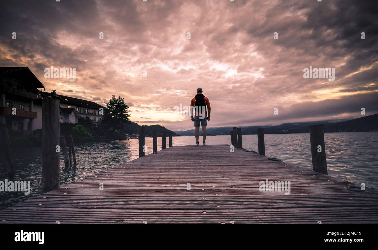 Uomo solitario in piedi sul bordo del molo di legno che guarda al lago calmo e la foresta dall'altro lato Foto Stock
