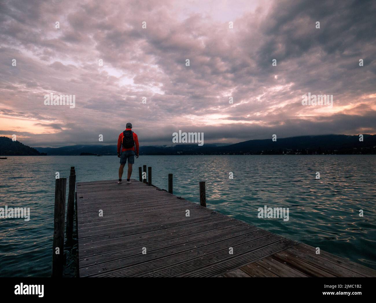Uomo solitario in piedi sul bordo del molo di legno che guarda al lago calmo e la foresta dall'altro lato Foto Stock