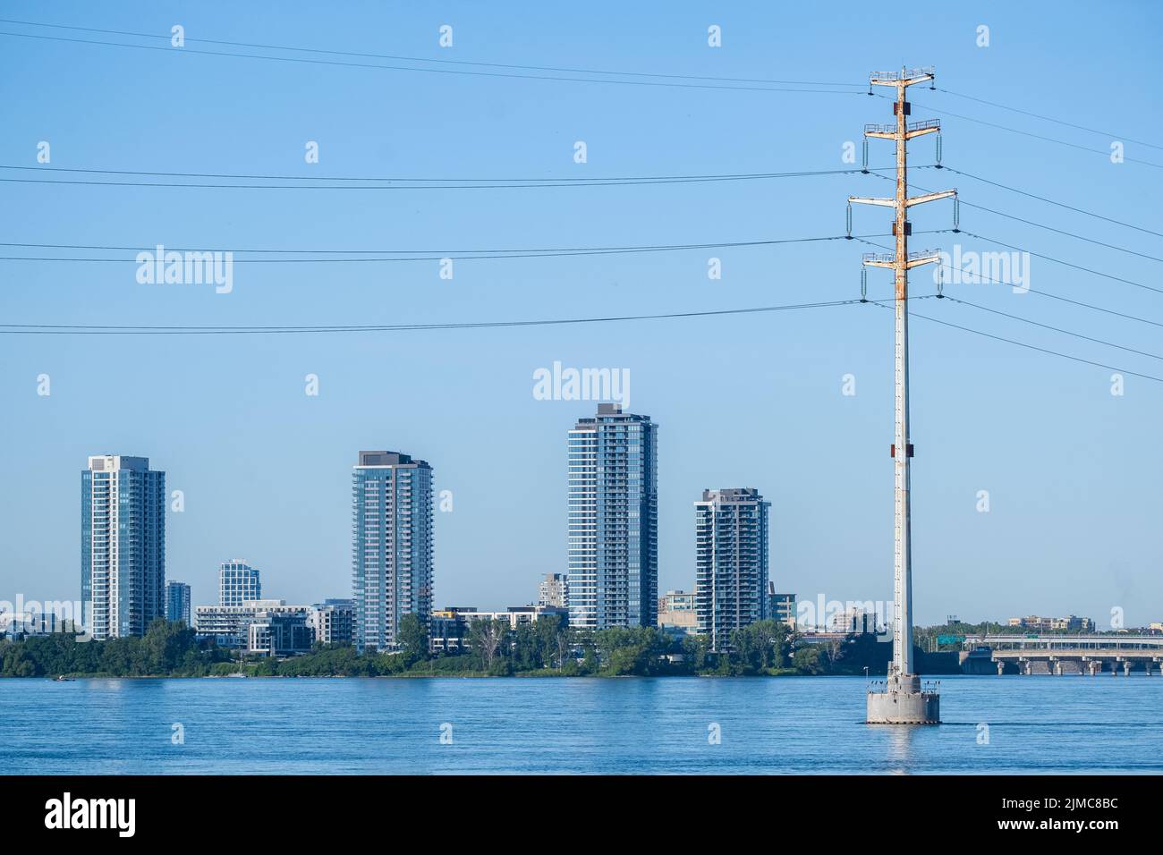 Un palo di distribuzione di energia elettrica si trova in un fiume con alti edifici sulla riva in lontananza. Foto Stock