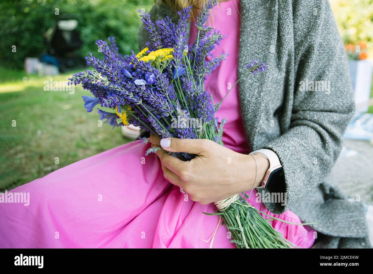 bouquet estivo di yarrow giallo e bouquet di lavanda nelle mani della donna, donna che indossa un abito rosa seduto in un parco Foto Stock