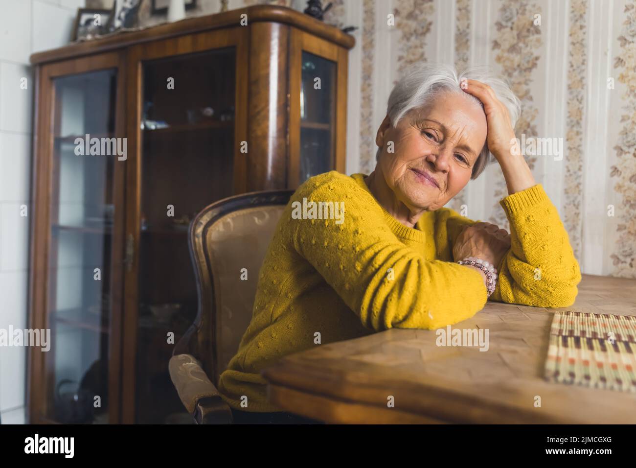 Bella piccola donna anziana dai capelli corti seduta su una sedia al tavolo del soggiorno in un piacevole umore con la testa appoggiata sul braccio e guardando via pensando al suo passato. Foto di alta qualità Foto Stock