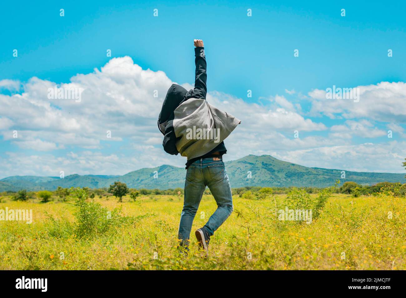 Uomo dalla parte posteriore che salta in un campo piacevole, vista posteriore dell'uomo che salta nell'erba che solleva il pugno, concetto di una persona libera che salta e solleva il suo braccio, libero Foto Stock