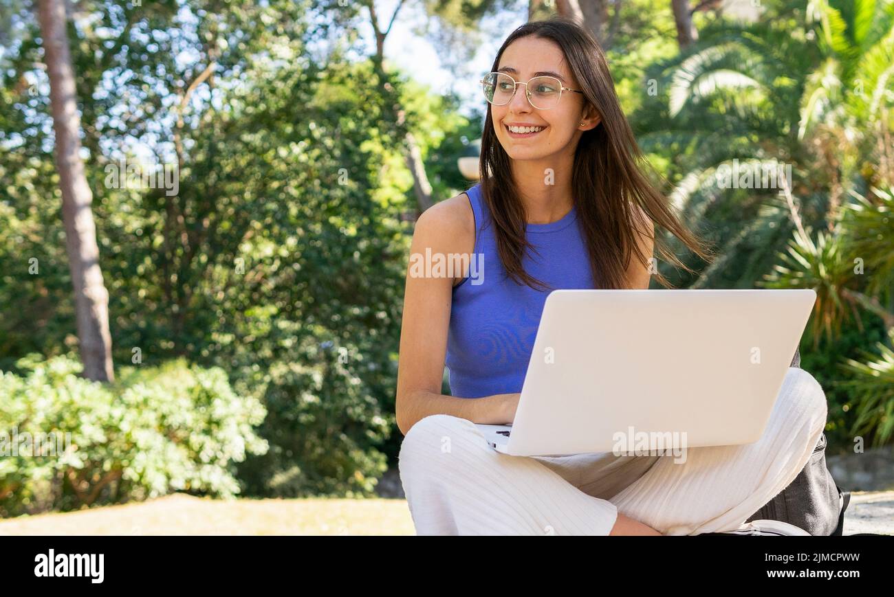 Studentessa femminile focalizzata a scrivere su netbook mentre si fa lavoro a casa su una superficie di pietra nel parco con alberi verdi la giornata estiva guardando via Foto Stock