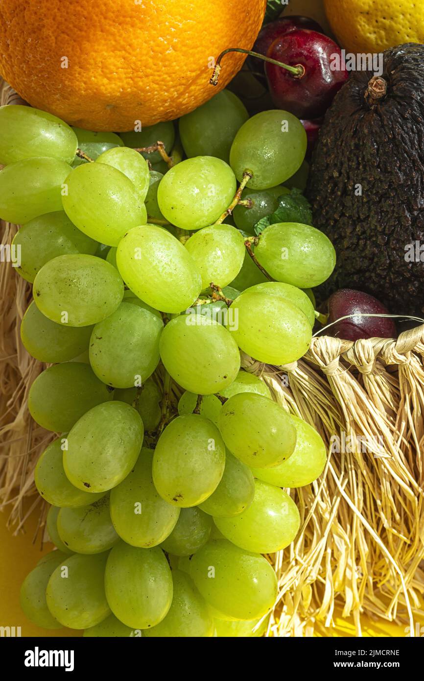 La natura morta di frutta e uva assortite è inserita in un cestino di vimini con foglie di menta verde sul tavolo su sfondo giallo e bianco con ombra Foto Stock