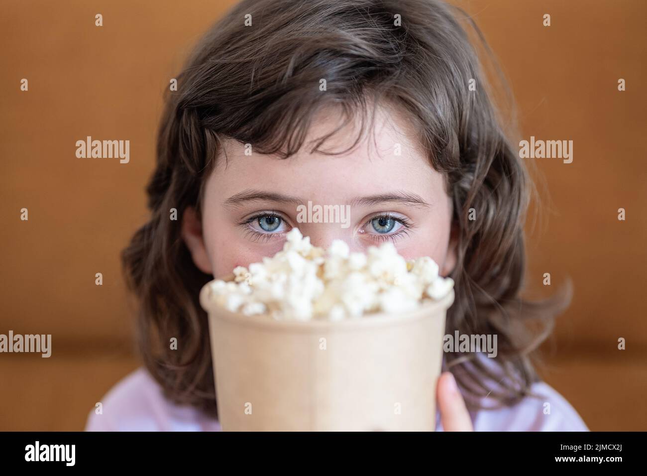 Ragazza positiva che guarda via mentre copre metà della faccia con secchio di cartone di popcorn delizioso sul divano durante lo spettacolo a casa guardando la macchina fotografica Foto Stock