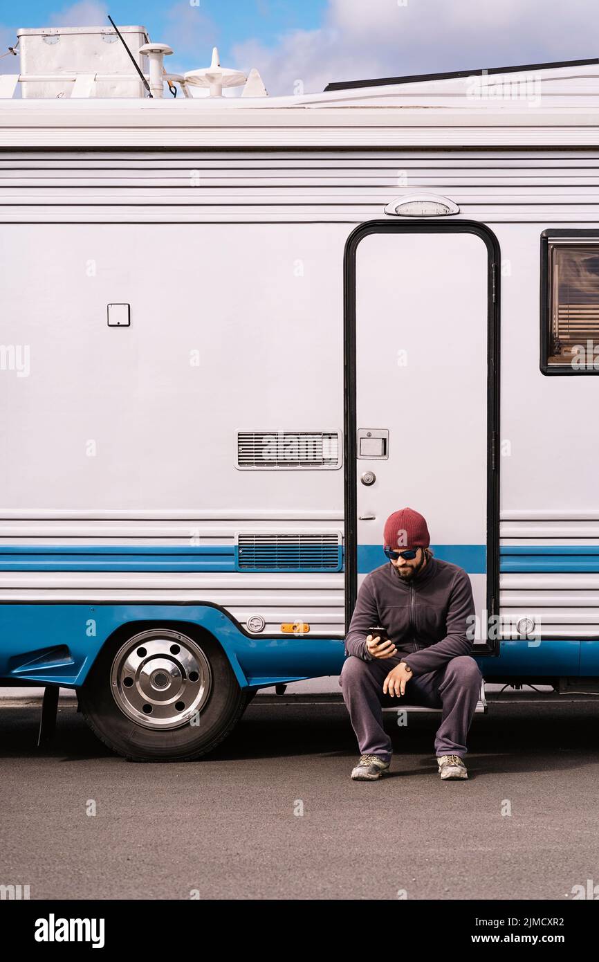 Pieno corpo di giovane viaggiatore con bearded maschile in abiti casual e occhiali da sole seduti sul gradino di camper e messaggi sullo smartphone Foto Stock