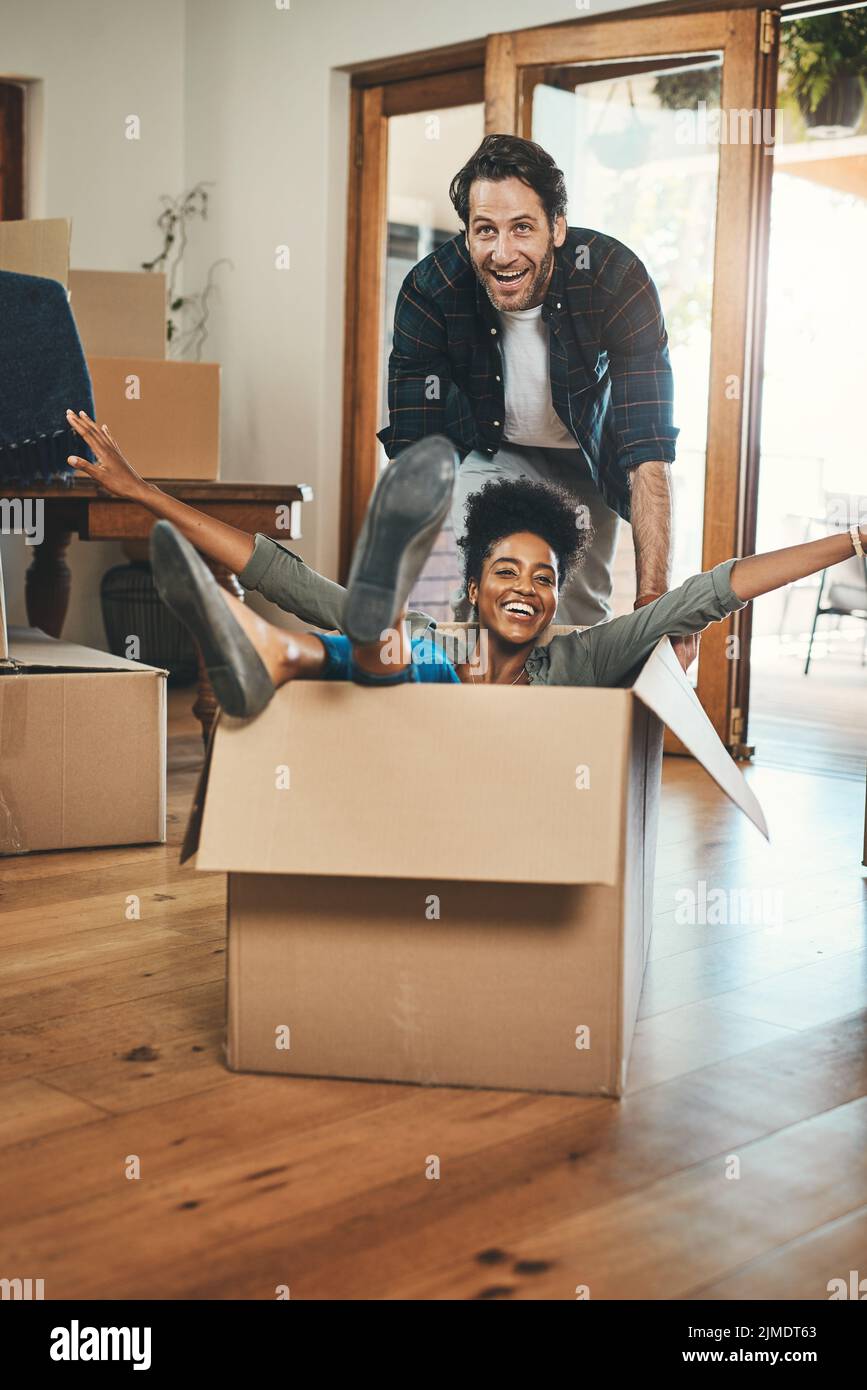 Divertimento, giocoso e ridendo proprietari di case giocando in scatola e godendo nuova sede come investitori immobiliari. Sciocco coppia interrazziale e uomo felice spingendo Foto Stock