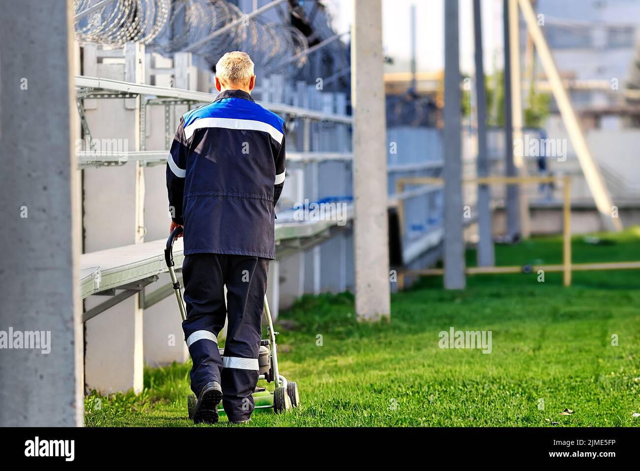 Un uomo anziano in abiti da lavoro cammina con un rasaerba in una giornata di sole. Un pensionato lavora a tempo parziale. Vista posteriore Foto Stock