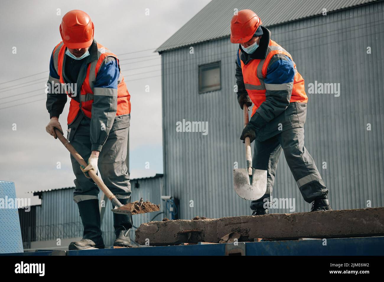 Due lavoratori in cappelli duri, abiti da lavoro e una maschera medica lavorano con pale in un cantiere. Duro lavoro fisico. Foto Stock