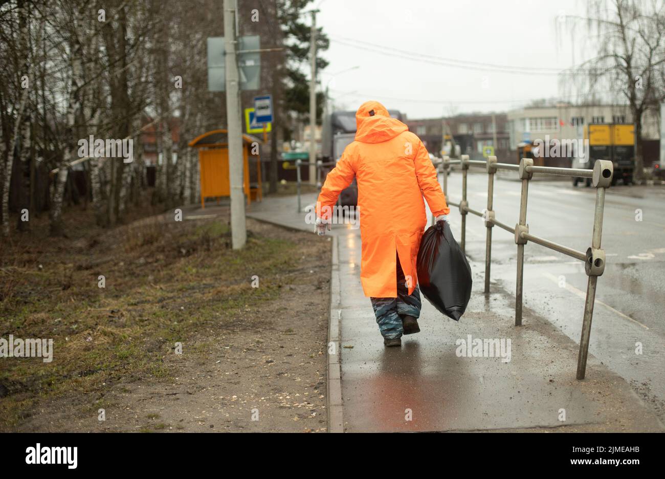 Il lavoratore rimuove i rifiuti dalla strada. Uomo sotto la pioggia d'arancio. Borsa nera in mano. Lavoratore su strada. Foto Stock