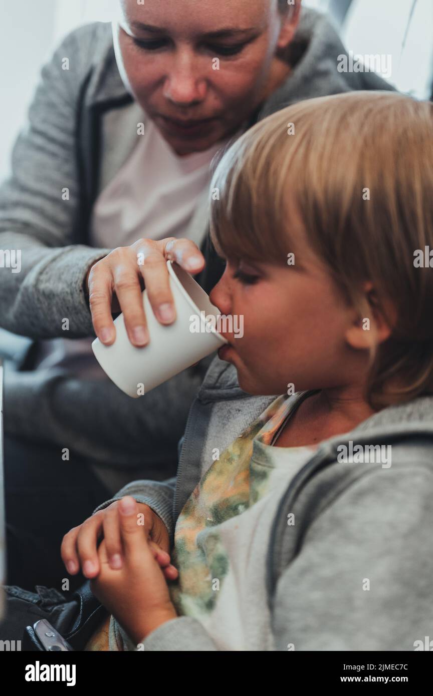 la mamma beve il ragazzino candido di cinque anni da una tazza di carta seduta sul sedile dell'aeroplano durante il volo. madre e bambini o bambino dentro Foto Stock